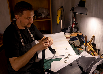 Artesão em sua oficina trabalhando com corte e preparação de tiras de couro sobre uma bancada com moldes de papel.